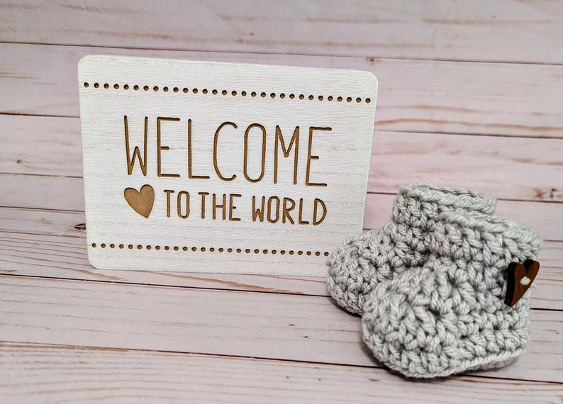 May include: A white wooden sign with the text "Welcome to the world" and a small heart.  A pair of gray crocheted baby booties with a wooden heart button.