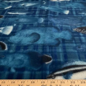 May include: A blue fleece fabric featuring a repeating pattern of various fish in shades of dark blue, light blue, and white. A wooden ruler is positioned at the bottom edge, showing measurements from 10 inches to 27 inches across the fabric.