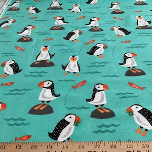 May include: A teal fabric featuring a repeating pattern of black and white puffins with orange beaks and feet, some standing on grey rocks or holding orange fish. A wooden ruler at the bottom shows measurements in inches from 9 to 23 inches.