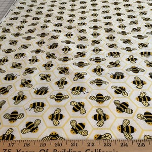 May include: A white fabric featuring a repeating pattern of black and yellow bees centered within light yellow hexagonal honeycomb outlines. A wooden ruler is visible at the bottom edge, showing measurements in inches from 14 to 28 inches.