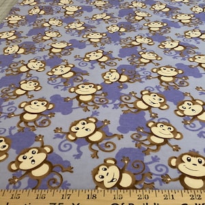 May include: Light blue fabric with a repeating pattern of cartoon monkeys in brown and cream colors. The monkeys are set against a background of purple monkey-shaped shadows. The fabric is measured in inches.