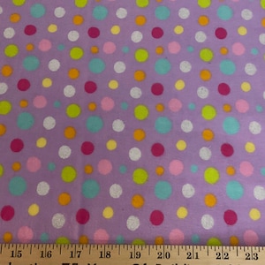 May include: A lavender fabric with a repeating pattern of colorful polka dots in shades of pink, green, blue, yellow, and white. The fabric is measured with a wooden ruler, showing inches.