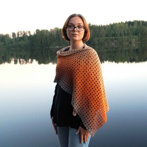 May include: A woman wearing a black shirt and blue jeans stands in front of a lake. She is wearing a crocheted shawl with a gradient of brown and orange.
