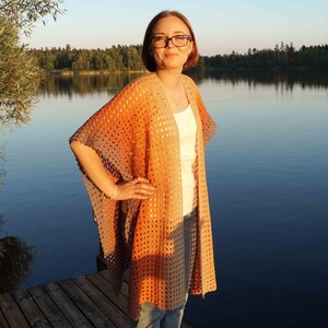Peut inclure: Une femme portant un châle en crochet avec un dégradé de couleurs orange et gris. Le châle est ouvert à l'avant et a une coupe ample. La femme porte un débardeur blanc et un jean bleu. Elle se tient sur une jetée en bois au bord d'un lac.