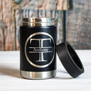 May include: Black stainless steel can cooler with a silver monogrammed circle featuring the letter "T" and the name "Anthony". The cooler has a black lid.