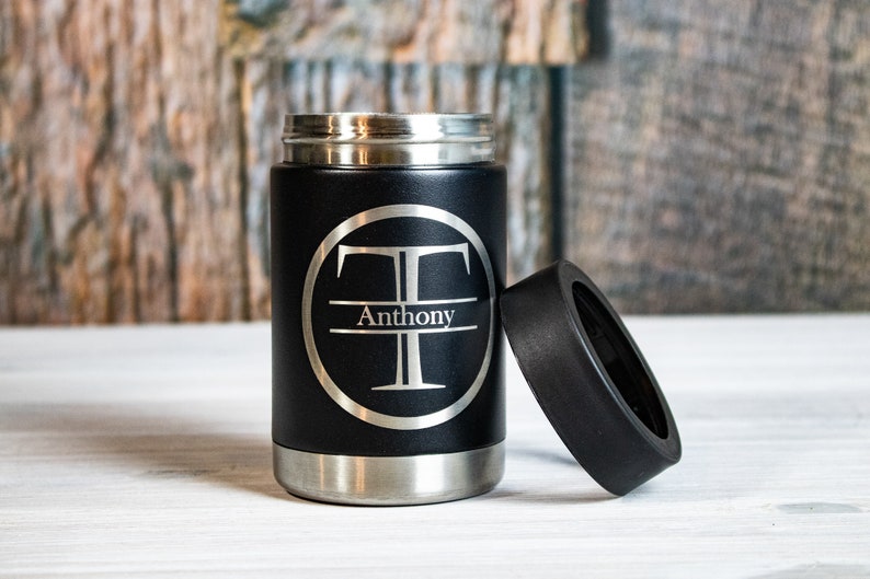 May include: Black stainless steel can cooler with a silver monogrammed circle featuring the letter "T" and the name "Anthony". The cooler has a black lid.