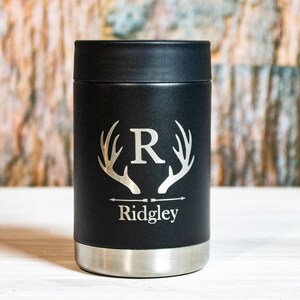May include: Black insulated can cooler with a silver bottom. The cooler has a deer antler design with the letter "R" in the center and the name "Ridgley" below.