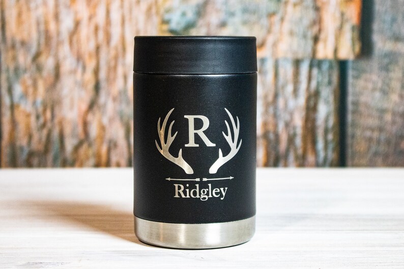 May include: Black insulated can cooler with a silver bottom. The cooler has a deer antler design with the letter "R" in the center and the name "Ridgley" below.