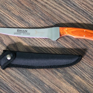 May include: A personalized knife with a wooden handle and a black sheath. The blade is engraved with the name "BRIAN" and the text "Groomsman - 10.10.2020".