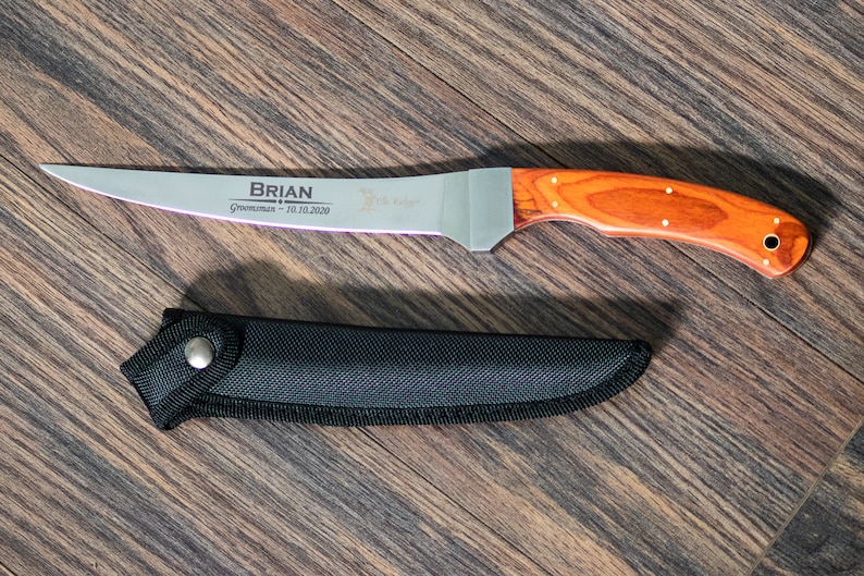 May include: A personalized knife with a wooden handle and a black sheath. The blade is engraved with the name "BRIAN" and the text "Groomsman - 10.10.2020".