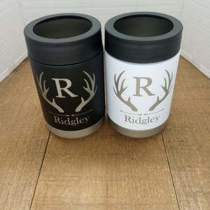 Op de afbeelding: Twee gepersonaliseerde roestvrijstalen blikjeskoelers met een zwarte en witte afwerking. Elke koeler heeft een hertengewei-ontwerp met de letter "R" en de naam "Ridgley" gegraveerd op de bodem.
