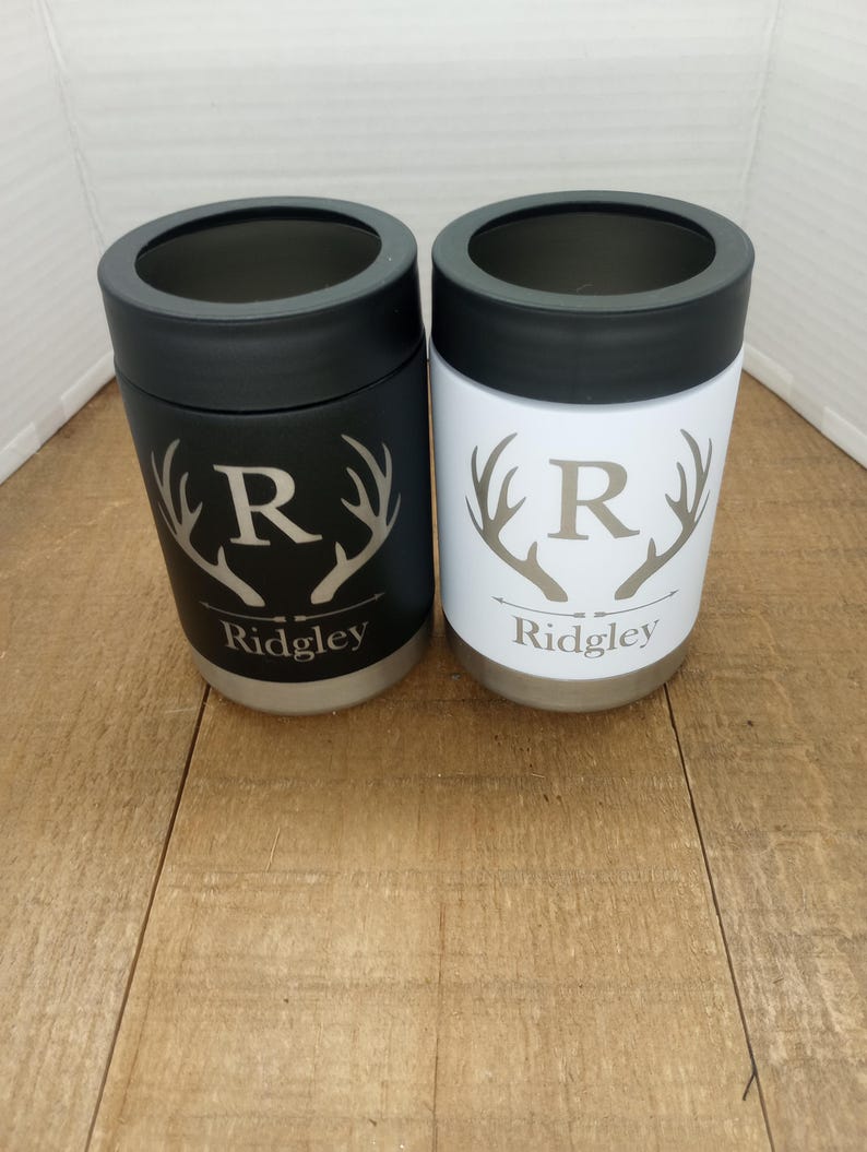 Op de afbeelding: Twee gepersonaliseerde roestvrijstalen blikjeskoelers met een zwarte en witte afwerking. Elke koeler heeft een hertengewei-ontwerp met de letter "R" en de naam "Ridgley" gegraveerd op de bodem.