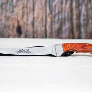 May include: A personalized hunting knife with a red wood handle and a silver blade. The blade is engraved with the name "DAVID" and the text "Groom - 10.10.2020".