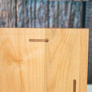 May include: The back of a light wood plaque with two keyhole slots for hanging. The wood has a natural grain pattern.