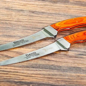 May include: Two personalized hunting knives with orange wood handles. The blades are engraved with the names "BRIAN" and "DAVID" and the date "10.10.2020".