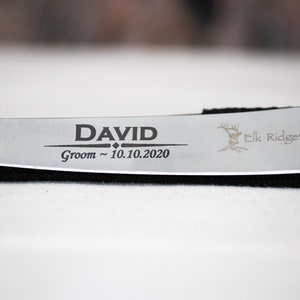 May include: A silver knife with a black handle engraved with the text "DAVID Groom ~ 10.10.2020" and a deer silhouette with the text "Elk Ridge".