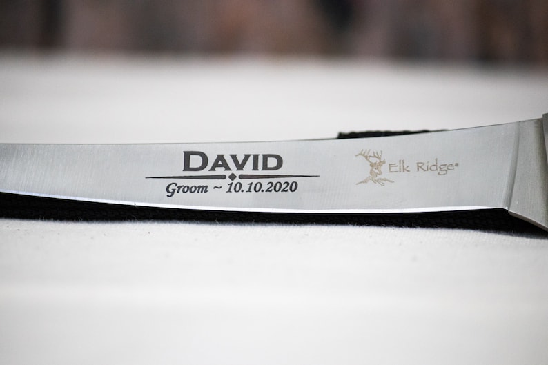 May include: A silver knife with a black handle engraved with the text "DAVID Groom ~ 10.10.2020" and a deer silhouette with the text "Elk Ridge".