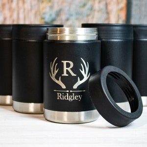 Op de afbeelding: Zes zwarte ge&iuml;soleerde blikjeskoelers met een zilveren band rond de bovenkant. E&eacute;n koeler heeft een gepersonaliseerd ontwerp met een monogram "R" in een gewei en de naam "Ridgley" eronder. De koeler is open en het deksel ligt ernaast.