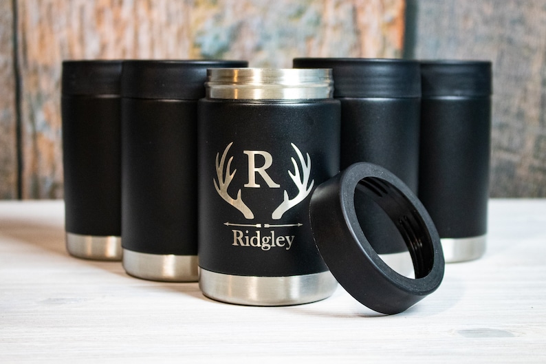 Op de afbeelding: Zes zwarte ge&iuml;soleerde blikjeskoelers met een zilveren band rond de bovenkant. E&eacute;n koeler heeft een gepersonaliseerd ontwerp met een monogram "R" in een gewei en de naam "Ridgley" eronder. De koeler is open en het deksel ligt ernaast.