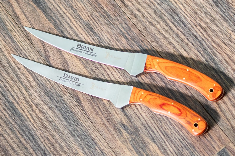 May include: Two personalized steak knives with wooden handles. The blades are engraved with the names "BRIAN" and "DAVID" along with the titles "Groom" and "Groomsman" and the date "10.10.2020".