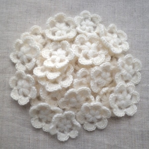 May include: A collection of small, off-white crochet flowers. Each flower has a five-petal design with a textured center. The flowers are arranged in a loose pile, creating a soft, handmade aesthetic. The background is a neutral, textured fabric.