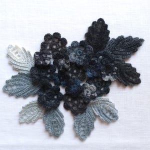 Könnte beinhalten: Eine Häkelapplikation mit einer Ansammlung von dunkelanthrazitfarbenen und schwarzen Blüten, umgeben von hellgrauen und weißen Blättern. Das Blumenarrangement ist mit komplexen Garnstichen gefertigt und zeigt einen Verlauf aus monochromatischen Grautönen.
