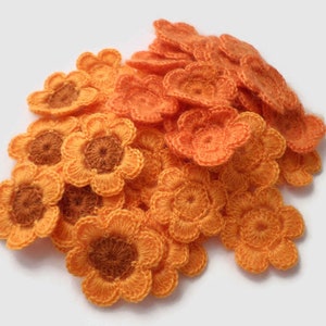 Puede incluir: Un montón de flores de ganchillo de color naranja y marrón. Las flores están hechas de hilo y tienen un aspecto delicado y hecho a mano.