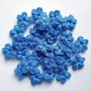 May include: A collection of small, five-petaled blue flowers crafted from yarn. Each individual flower features a textured, crocheted design with a central point and rounded petals, arranged in a loose cluster against a plain white background.