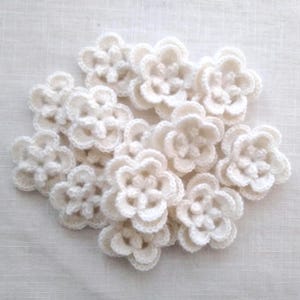 May include: A collection of off-white, crocheted flower appliques. Each flower has multiple layers of petals, creating a three-dimensional effect. The flowers are clustered together, showcasing their delicate texture and handmade quality.