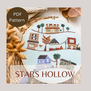 May include: A cross-stitch embroidery hoop with a blue background and colorful houses, trees, and lights. The text "STARS HOLLOW" is stitched in brown thread at the bottom of the hoop.