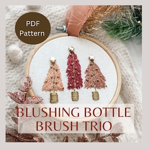 May include: A PDF pattern for embroidery featuring three small Christmas trees in a hoop. The trees are embroidered in shades of pink, red, and gold. The text "BLUSHING BOTTLE BRUSH TRIO" is below the hoop.