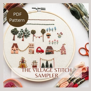 Könnte beinhalten: Ein weißer Stickrahmen mit einer bunten Stickerei einer Dorfszene. Die Szene enthält Häuser, Bäume, ein Auto und Blumen. Der Text "THE VILLAGE STITCH SAMPLER" steht unter der Szene.