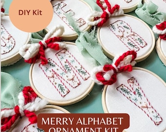 May include: A DIY kit for creating embroidered alphabet ornaments. Each ornament features a letter, with festive designs in red, green, and white. The text "MERRY ALPHABET ORNAMENT KIT" is displayed on a brown banner.