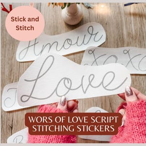 May include: White fabric stickers with grey script lettering spelling "Love", "Amour", and "XOXO". The stickers are labelled "Stick and Stitch" and "Words of Love Script Stitching Stickers". The background is a wooden surface.