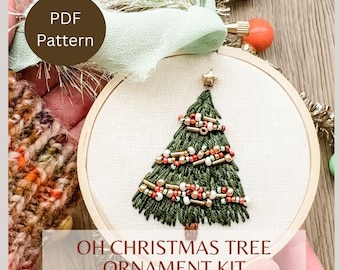 May include: A DIY Christmas tree ornament kit featuring a wooden embroidery hoop with a green, red, and gold beaded Christmas tree design. The text "OH CHRISTMAS TREE ORNAMENT KIT" is printed below the hoop.