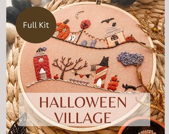 Puede incluir: Un bastidor de bordado con un diseño de pueblo de Halloween. El diseño incluye casas bordadas, calabazas, un gato negro y un árbol espeluznante. También se ve el texto "Full Kit" y "Halloween Village".