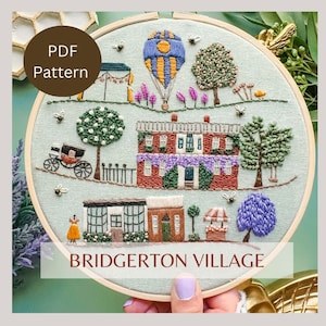 May include: A hand-stitched embroidery hoop with a colorful scene of a village with houses, trees, a hot air balloon, and the text "Bridgerton Village".