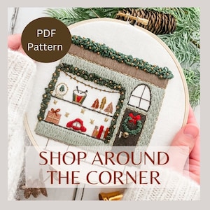May include: A PDF pattern for a hand-stitched embroidery hoop art piece depicting a small shop with a green awning, a red door, and a window display of Christmas decorations. The text "SHOP AROUND THE CORNER" is below the hoop.