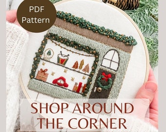 May include: A PDF pattern for a hand-stitched embroidery hoop art piece depicting a small shop with a green awning, a red door, and a window display of Christmas decorations. The text "SHOP AROUND THE CORNER" is below the hoop.
