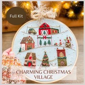 May include: A hand-stitched embroidery hoop featuring a charming Christmas village scene. The design includes houses, a Christmas tree, and a red barn, all set against a light blue background. The text "Charming Christmas Village" is displayed at the bottom.