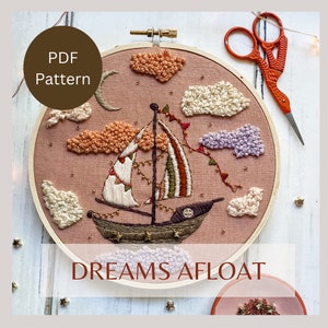 May include: Embroidered hoop art featuring a sailboat with flags and lights sailing through a pink sky with fluffy white and purple clouds. The hoop is surrounded by gold glitter stars and the text "DREAMS AFLOAT" is printed below the hoop.