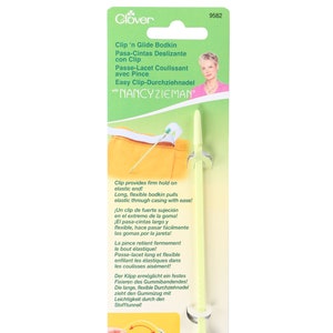 May include: Green and white plastic bodkin with a clip on the end. The bodkin is designed to help thread elastic through casings. The packaging includes instructions in English, French, German, Spanish, and Italian. The product is by Clover and features the name Nancy Zieman.