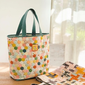 May include: A quilted tote bag featuring a colorful hexagonal patchwork pattern in various pastel and bright tones, with dark green polka-dot handles. Beside it lies a second quilted bag with a rectangular grid pattern in orange, pink, and grey.