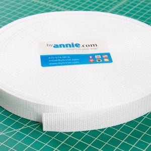 May include: A roll of white webbing material, likely for crafting or sewing projects, sits on a green cutting mat. A label with the text "byannie.com" and contact information is visible on the webbing.