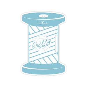 May include: A light blue spool-shaped sticker with the text "Quilty Tools" in a circular design. The sticker has a white and blue striped pattern and the words "Riley Blake Designs" are printed on the top.
