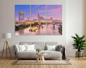 May include: A three-panel wall art set depicting the Nashville skyline at dusk. The image features a river with a paddlewheel boat, a steel truss bridge, and city skyscrapers under a vibrant pink and blue sky, illuminated by warm city lights.