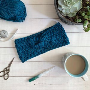 May include: A teal blue crocheted headband with a knotted detail. The headband is on a white wooden surface with a ball of yarn, a measuring tape, a crochet hook, a cup of coffee, and a potted succulent.