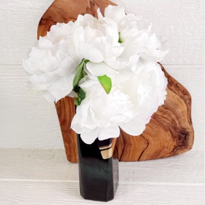 May include: A bouquet of white peonies in a dark green and gold vase. The vase is sitting on a wooden cutting board.