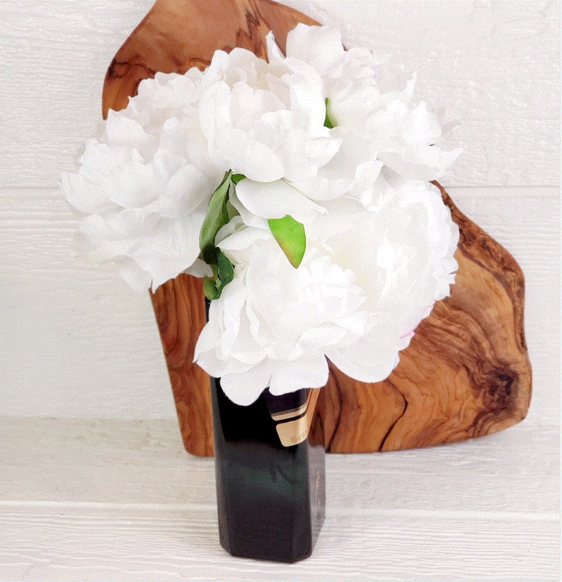 May include: A bouquet of white peonies in a dark green and gold vase. The vase is sitting on a wooden cutting board.