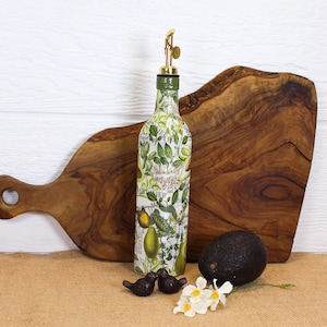 May include: A glass oil dispenser bottle decorated with a botanical print of green avocados, leaves, and text, topped with a gold-colored metal pour spout. The bottle is positioned in front of a wooden cutting board with a dark brown avocado nearby.